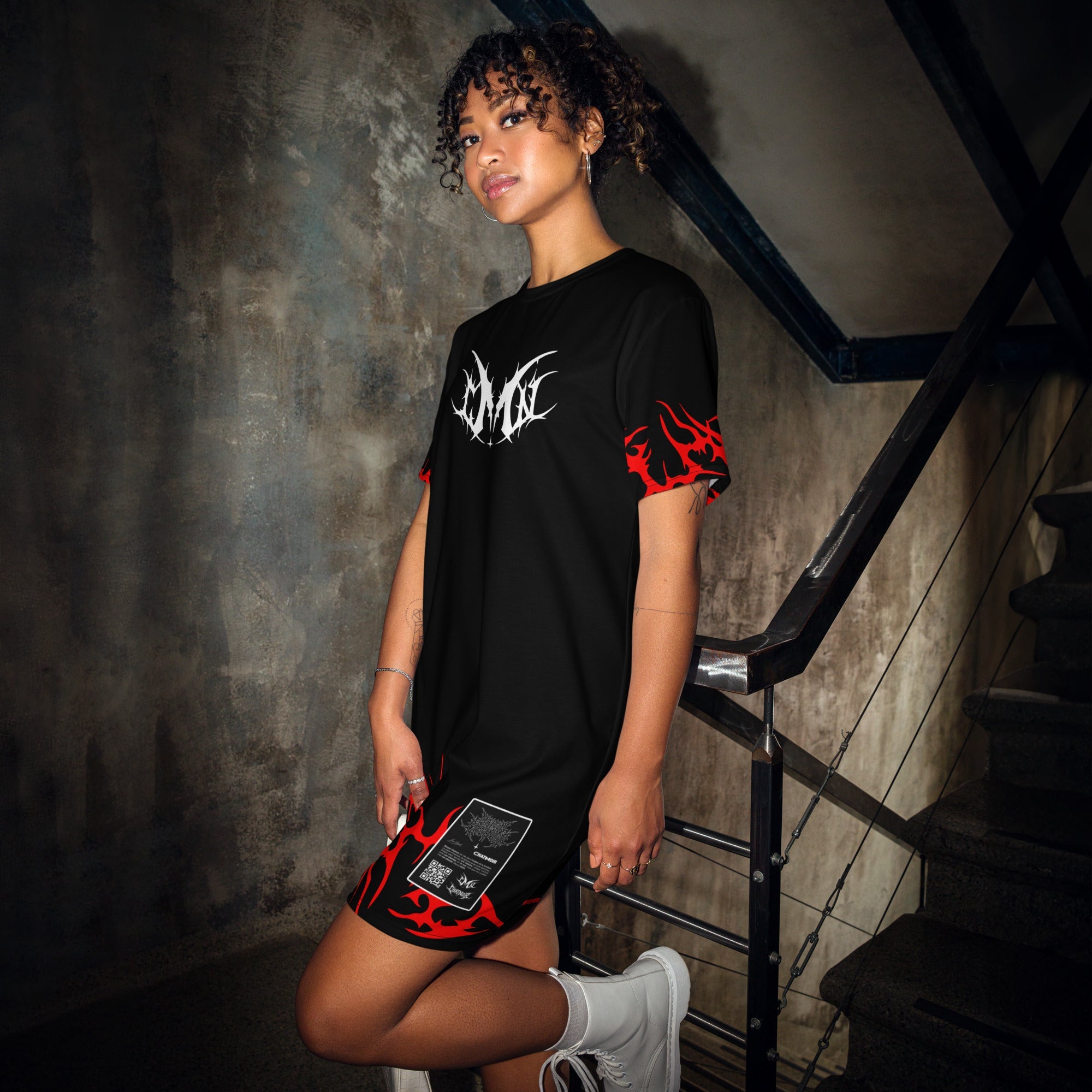 Femme posant dans un escalier urbain avec la robe t-shirt Maison Chatnoir noire, affichant un logo griffé MCN blanc et des motifs de flammes tribales rouges.