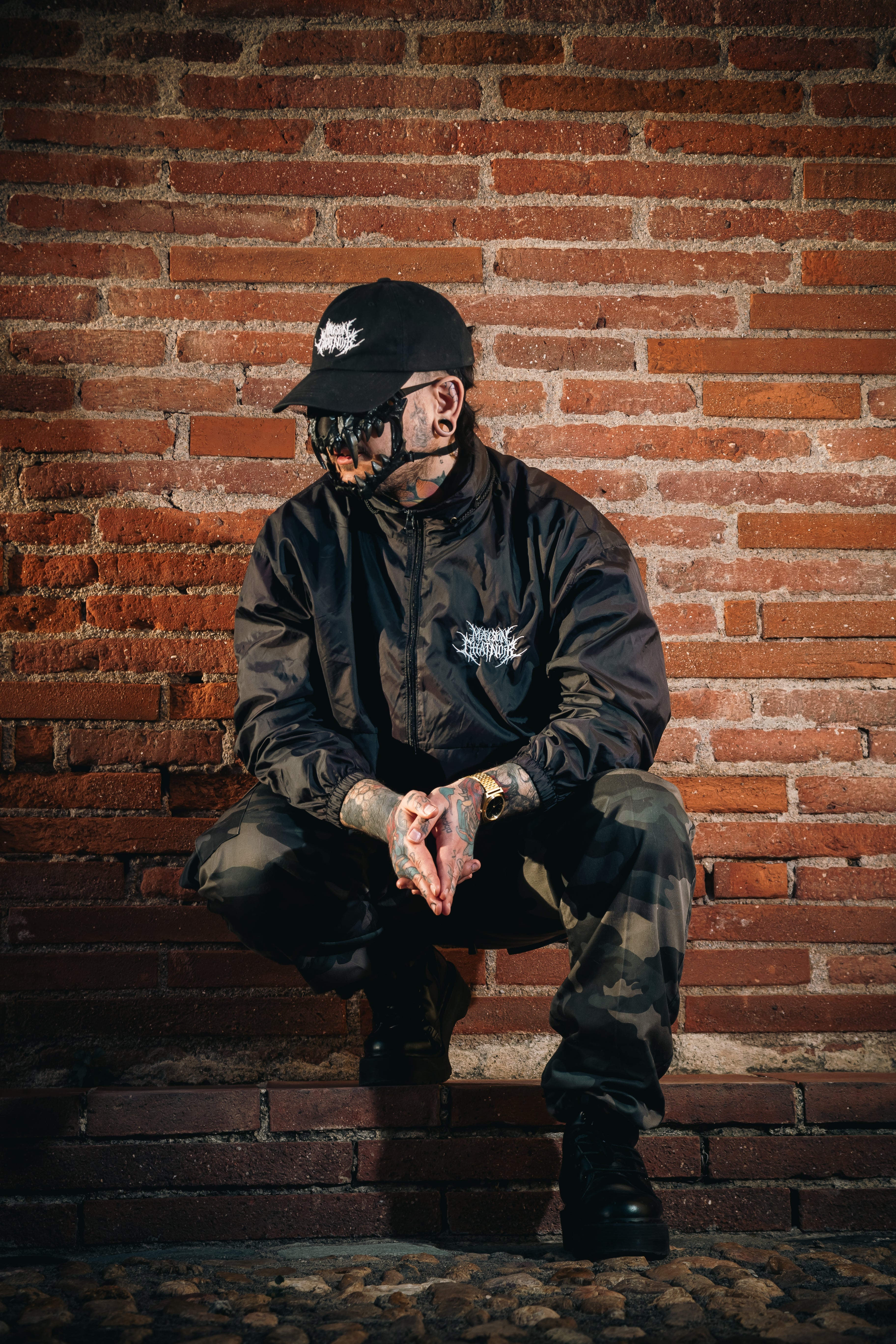 Homme accroupi devant un mur de brique rouge, masque de chat sur le visage, casquette et parka noires avec logo Maison ChatNoir Blanc