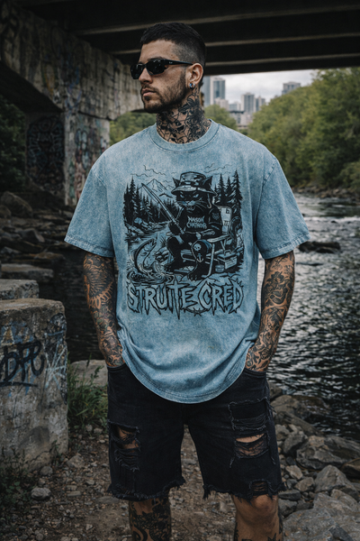 Homme tatoué portant le t-shirt bleu effet Snow Wash Maison Chatnoir, design exclusif StruiteCred Fishing Cat. Coupe oversized et style streetwear radical sous un pont de graffitis au bord d'une rivière.